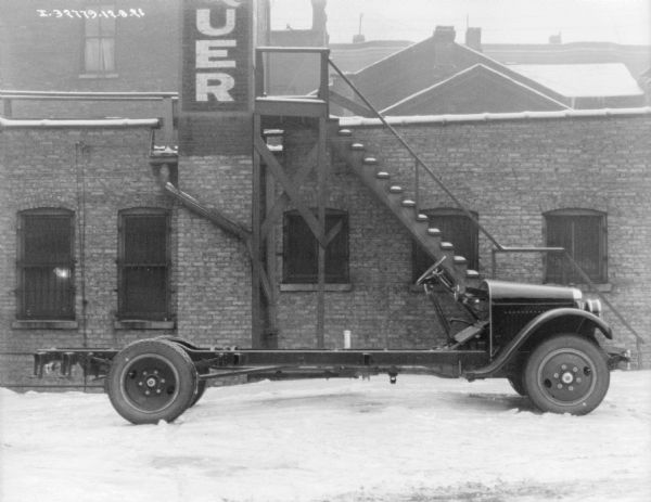 Right side profile view of a truck parked outdoors in the snow. The truck has fenders and a front hood, but the chassis is exposed. A brick building is in the background.