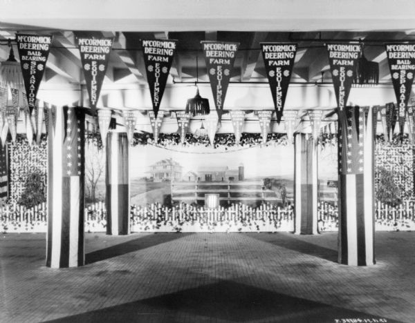 View of indoor exhibition display with farm scene on the back wall. Hanging along the ceiling are pennants that read: "McCormick-Deering Ball Bearing Separators," and McCormick-Deering Farm Equipment." U.S. flags are draped on the columns of the building.