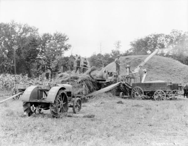 View across field towards men working with a tractor belt-driving a thresher. Two men are standing on the back of a hay wagon loading a thresher. Another man in the center is standing on the thresher, which is blowing hay onto a large haystack in the background. Another man is standing near the chute to which is filling up a horse-drawn wagon with grain.