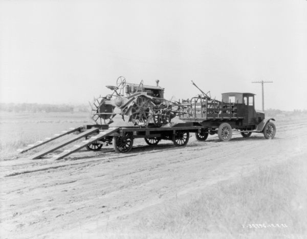 View down road towards a Farmall tractor loaded on a trailer, with another farm implement. A truck with stake body is pulling the trailer, and in the truck bed is another implement.