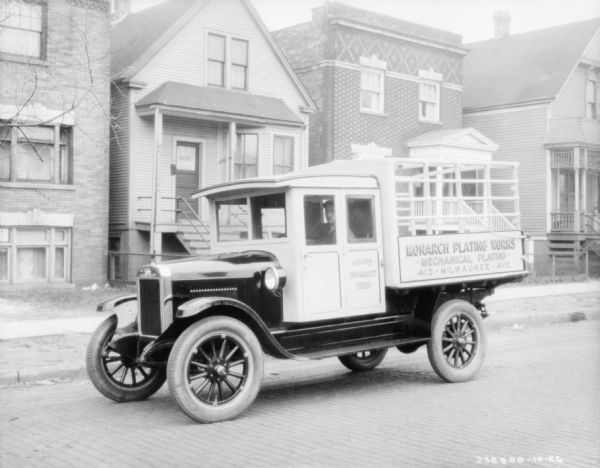 View across street towards a man sitting in the driver's seat of a delivery truck. The sign painted on the side of the truck reads: "Monarch Plating Works." There are wood and brick buildings sitting close together along the sidewalk in the background. Location: Haymarket.