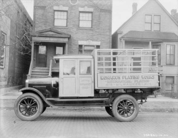 View across street towards a delivery truck parked along a curb. The sign painted on the side of the truck reads: "Monarch Plating Works." There are wood and brick buildings sitting close together along the sidewalk in the background. Location: Haymarket.