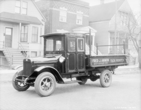 View across street towards a man sitting in the driver's seat of a delivery truck. The sign painted on the side of the truck reads: "Fred J. Ammon, Tuck Pointer." There are wood and brick buildings sitting close together along the sidewalk in the background. Location: Haymarket.