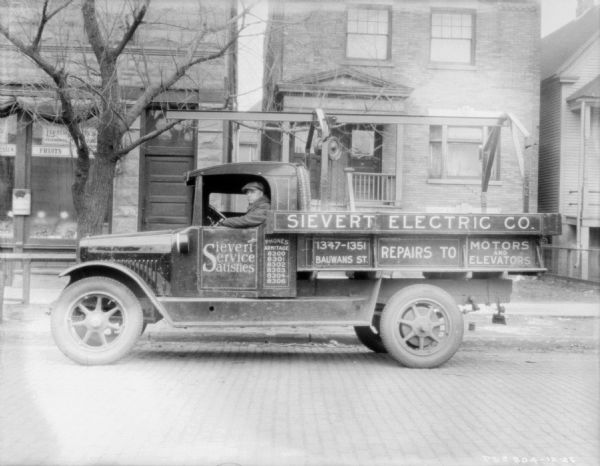 View across street towards a man sitting in the driver's seat of a delivery truck. The sign painted on the side of the truck reads: "Sievert Service Satisfies," and "Sievert Electric." There are wood and brick buildings sitting close together along the sidewalk in the background. Location: Haymarket.