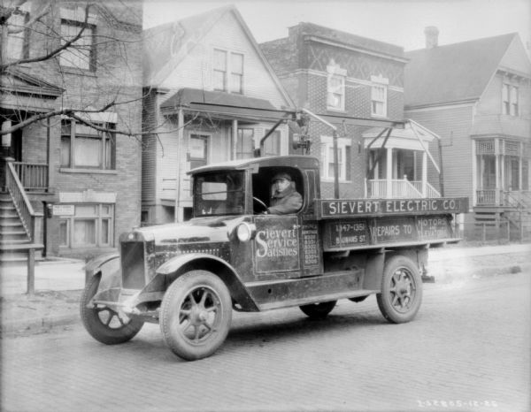 View across street towards a man sitting in the driver's seat of a delivery truck. The sign painted on the side of the truck reads: "Sievert Service Satisfies," and "Sievert Electric." There are wood and brick buildings sitting close together along the sidewalk in the background.