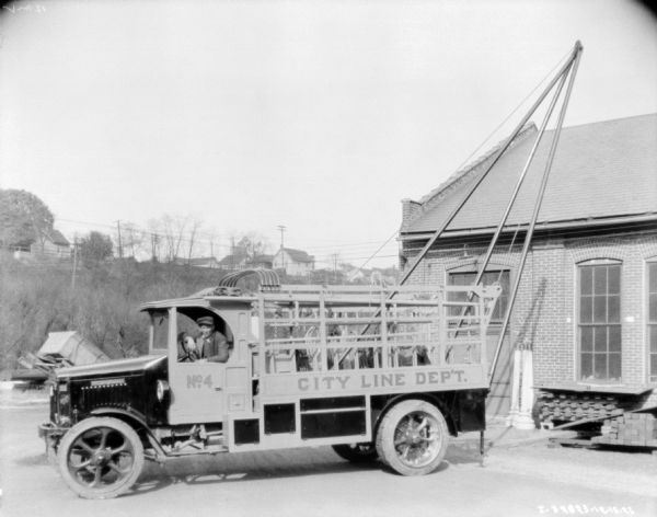 A man is sitting in the driver's seat of a truck. On the side of the truck is a sign that reads: "City Line Dep't." Equipment is hanging in the back of the truck which has a stake body. There is also a tall metal tripod attached with a cable suspended at an angle off the back of the truck.