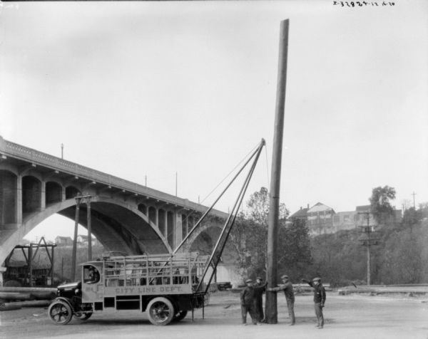 A man is in the driver's seat of a truck, helping a group of four men to erect a telephone pole, using a tall metal tripod attached to the back of the truck. On the side of the truck is a sign that reads: "City Line Dep't." Equipment is hanging in the back of the truck which has a stake body. There is a bridge in the background.
