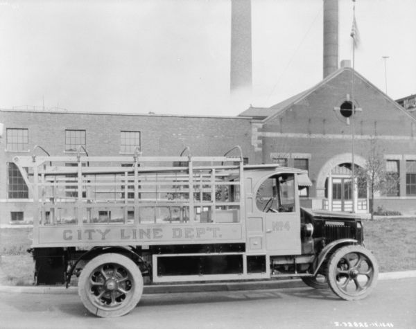 Right side of a truck with a sign that reads: "City Line Dep't." Equipment is hanging in the back of the truck which has a stake body. A large industrial building with large smokestacks is in the background. Above the arched entry to the building is a sign that reads, in part: "Richmond Municipal ___."