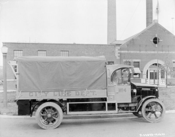 Right side of a truck with a sign that reads: "City Line Dep't." The back of the truck is covered with a tarp. A large industrial building with large smokestacks is in the background. Above the arched entry to the building is a sign that reads, in part: "Richmond Municipal ___."