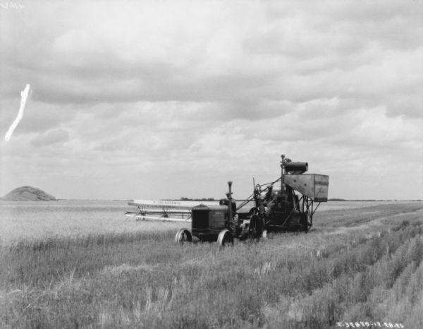 View across field towards a man driving a tractor in a field. Behind the tractor is a man sitting on the front of the harvester-thresher. There is a large pile of grain in the background on the left.