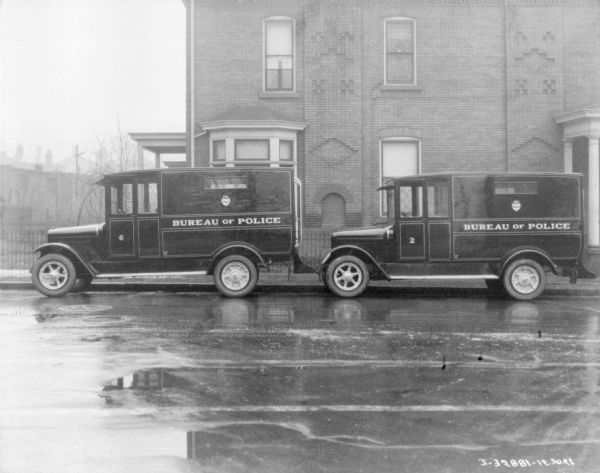 View across street towards two police trucks parked along the curb near a large brick building. The signs on the side of the trucks reads: "Bureau of Police."