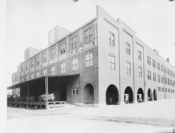 View across street towards the back and side of a large IHC dealership. There is a covered loading dock on the back, and vehicles are parked under archways on the side of the building. Painted signs on the brick wall above the loading dock read: "Mill__ Brothers Hardware Co. International Harvester Co. of America."