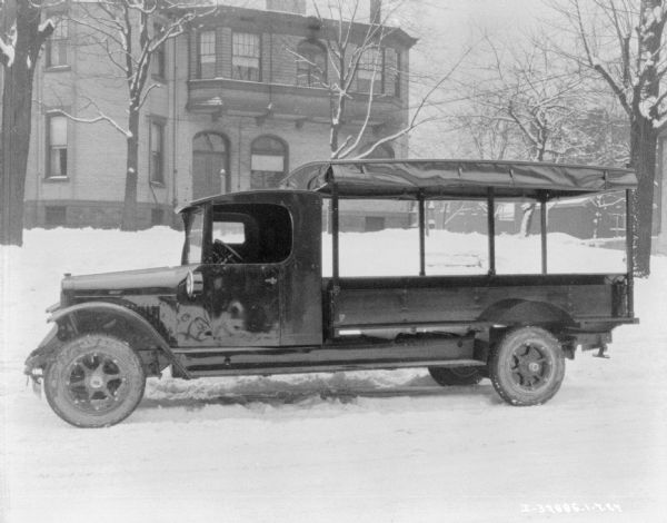 Left side profile view of truck/moving van parked on a snow street. The canvas sides of the bed of the truck are rolled up onto the roof. In the background is a large, brick building.