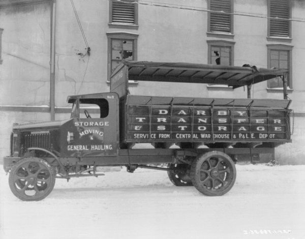 Left side view of a moving van parked in the snow.  The canvas sides of the bed of the truck are rolled up onto the roof. There is a large building with broken windows in the background. The sign painted on the side of the truck reads: "Darby Transfer & Storage."