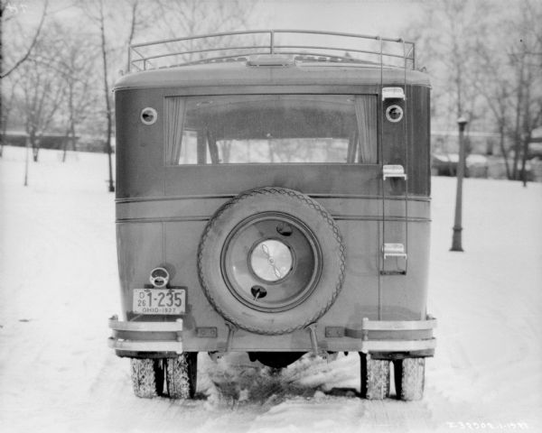 Rear view of International truck used as a bus. There is an Ohio license  plate on the back, and a spare tire. A small metal ladder is also on the back for access to the luggage rack on the roof. Snow is on the ground.