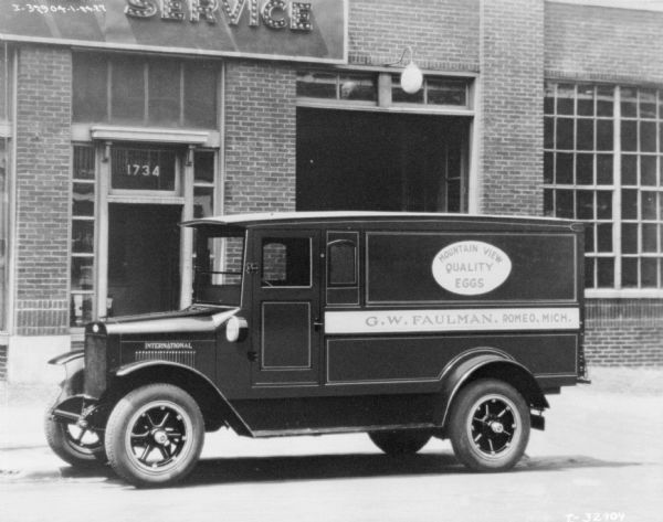 View across street towards a delivery truck parked along a curb. The sign painted on the back of the truck reads: "Mountain View Quality Eggs," and "G.W. Faulman." A brick building behind the truck has a sign above the entrance that reads: "Service."