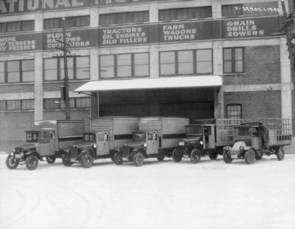 View across snowy yard towards delivery trucks backed up to a large loading dock on a multi-story brick building. Signs above the loading dock read: "Farm Wagon Trucks," "Tractors Oil Engines Silo Fillers" and "Plows, Harrows, Cultivators."