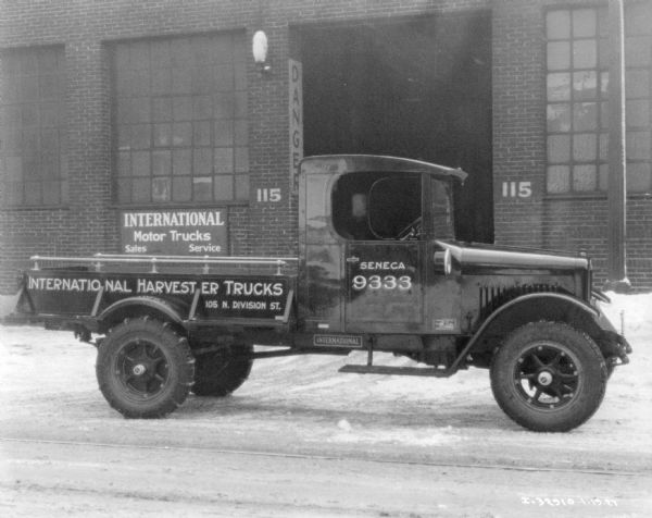 International  truck parked outdoors in front of a brick building with a large garage door. A sign on the building reads: "International Motor Trucks, Sales, Service." Snow is on the ground.