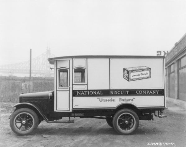 Left side profile view of a Uneeda Biscuit Company truck. There is a building on the right, and in the background is a bridge.