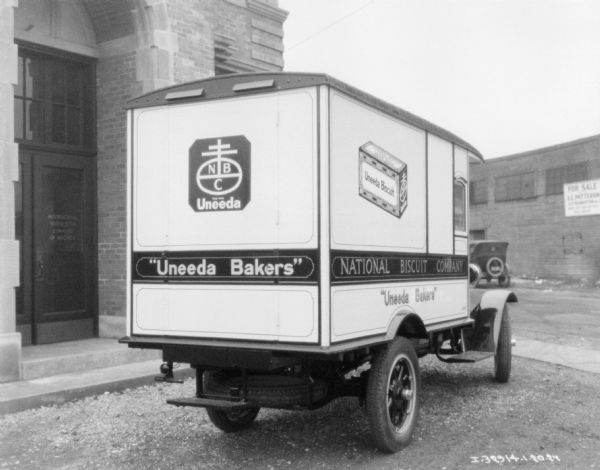 Three-quarter view from right rear of a Uneeda Bakers delivery truck, parked near the entrance to a brick building. The sign on the door of the building reads: "International Harvester Company of America."