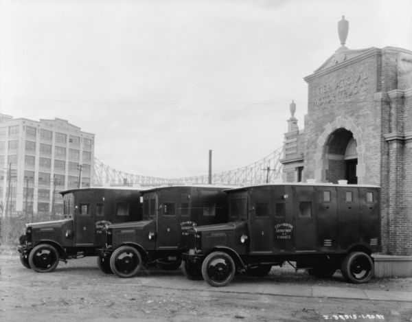 Armored trucks of department of finance for New York City near an IH plant. The trucks are lined up near a brick building, which has a sign that reads: "International Motor Trucks." There is a bridge in the background.