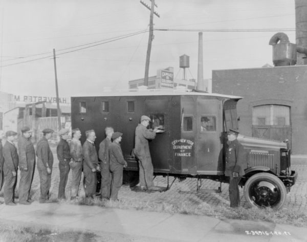 Men receiving paychecks from armored trucks parked outdoors at an IH plant. Signs on the trucks read: "Department of Finance, City of New York." A police officer is standing near the truck on the right.