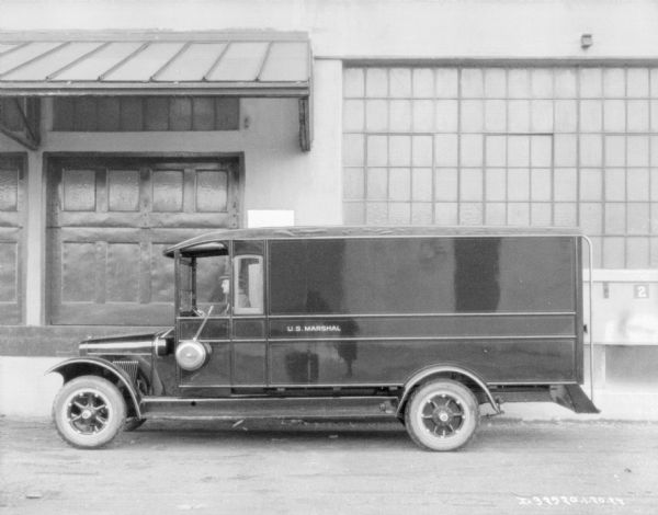 Driver's side view of a truck. A man is sitting in the driver's seat of the truck, and on the side is a painted sign that reads: "U.S. Marshall." The photographer is reflected on the side of the truck. There is an industrial building in the background, with a large awning over garage doors.