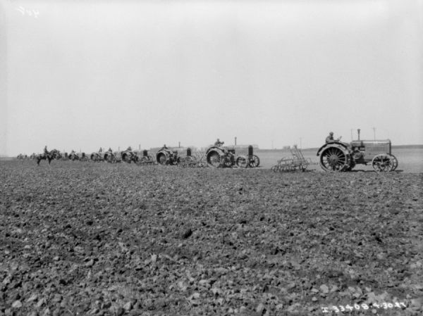 View across field towards ten or more men on tractors pulling cultivators. The men are lined up behind each other, and on the far left is a man on horseback. Farm buildings are in the far background.