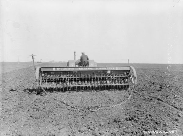Rear view of a man driving a tractor pulling a seeder. The sign on the seeder reads: "McCormick-Deering Power Lift 8. The number "7" is on the fenders over the wheels of the tractor.