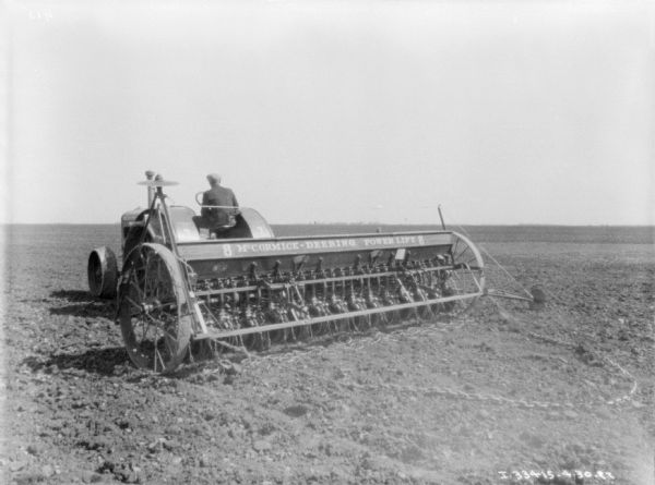 Three-quarter view from left rear of a man driving a tractor, pulling a seeder in a field. The sign painted on the back of the seeder reads: "McCormick-Deering Power Lift 8."