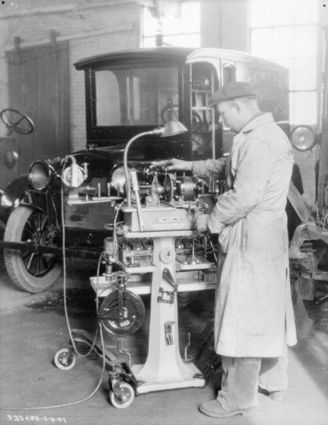 A man wearing a long jacket and a hat is working at a machine assembling parts for a truck, parked in the background.
