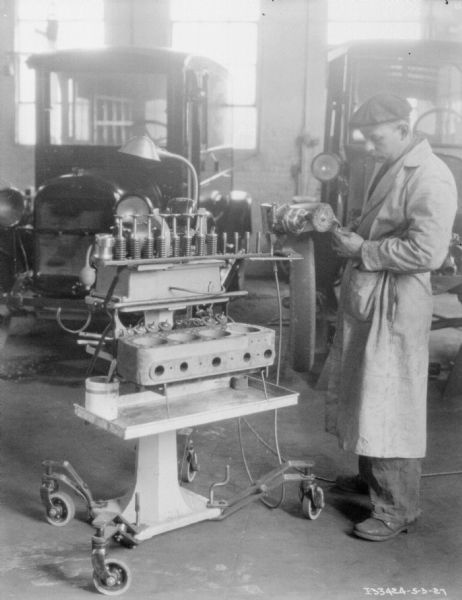 A man wearing a long jacket and a hat is working at a machine assembling parts for trucks parked in the background.