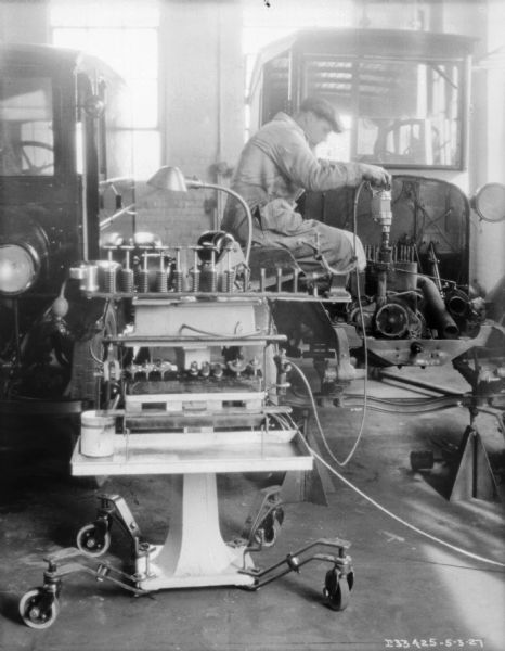 A man wearing a long jacket and a hat is working on the engine of a truck. A work cart is in the foreground.
