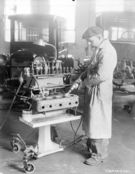 A man wearing a long jacket and a hat is working at a machine assembling parts for trucks parked in the background.