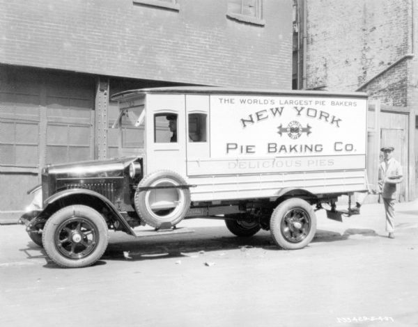 View across street towards a man sitting in the driver's seat of a delivery truck. A sign painted on the back of the truck reads: "New Yor Pie Baking Co." and "Delicious Pies." Another man is standing at the back of the truck holding pies in his arms. In the background are brick buildings. There is a spare tire mounted on a running board below the driver's side window.