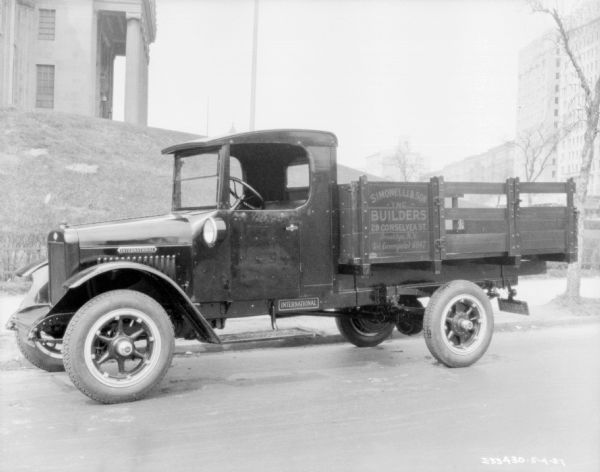 View across street towards an International truck. A sign on the side of the truck bed reads: "Simonelli & Son — Inc. — Builders." There is a large building with a columned entrance on a hill in the background on the left. On the right is a tall, commercial building.
