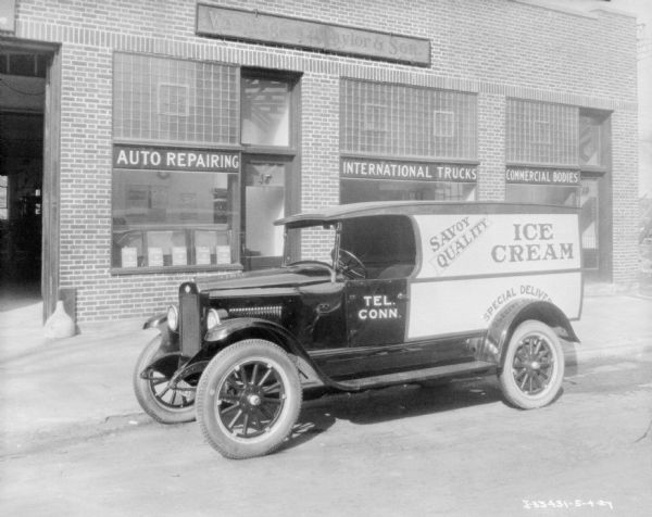 View across street towards a dairy delivery truck parked along a curb. The sign painted on the side of the truck reads: "Ice Cream, Savoy Quality." The building behind the truck has signs above show windows that read: "Auto Repairing," "International Trucks" and "Commercial Bodies." On the far left is an open garage door.