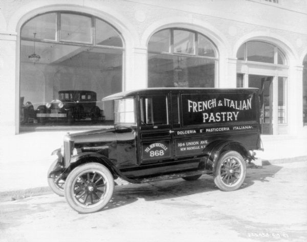 View across street towards a bakery delivery truck parked along a curb. A sign painted on the side of the truck reads: "French & Italian Pastry." A building in the background has large, arched windows, and inside is a showroom with a parked car on display.