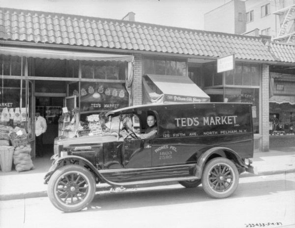 View across street towards a man sitting in the driver's seat of a grocery delivery truck. The sign on the side of the truck reads: "Ted's Market, 125 Fifth Ave., North Pelham, N.Y." The truck is parked in front of a storefront, which has a sign that reads: "Ted's Market." A man wearing a white jacket is standing just inside the doorway. Bushel baskets, boxes, and sacks hold vegetables on display on the sidewalk in front of the show windows.