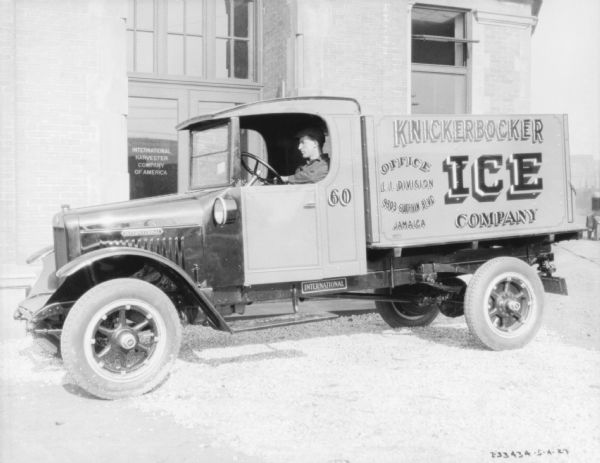 View across street towards a man sitting in the driver's seat of an ice delivery truck parked in front of a brick building. The sign on the door of the building reads: "International Harvester Company of America." The sign on the side of the truck bed reads: "Knickerbocker Ice Company."
