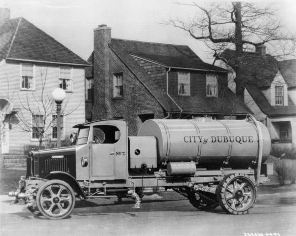 View across street towards a "City of Dubuque" oil delivery truck parked along the curb in a residential neighborhood. Three houses are in the background.