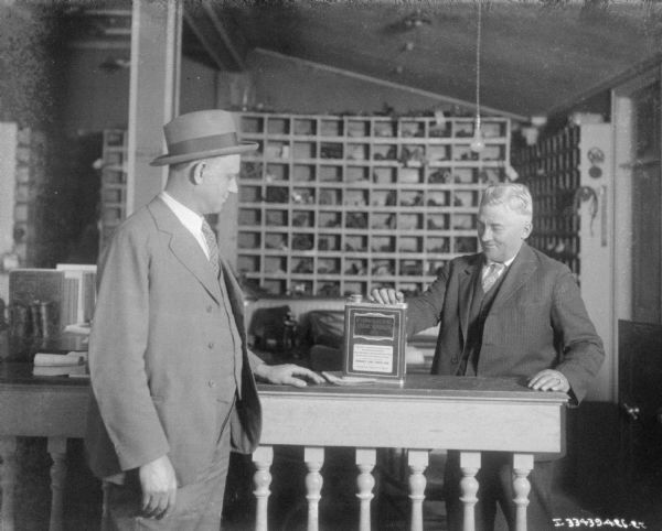 Two men are standing inside a dealership. One man is standing with his hand on a metal can on a counter with a label that reads: "McCormick-Deering Cream Separator Oil."