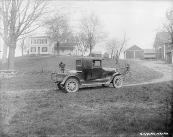 View uphill towards a truck on a driveway with a cream separator in the bed of the truck. A sign painted on the side of the open truck bed reads: "McCormick-Deering, Power Farm Equipment." A farmhouse, barn, and other farm buildings are in the background further up the hill.