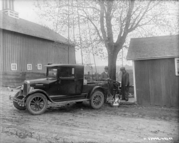 Two men are standing at the back of an International truck. There is a cream separator set up next to a farm building. There is a barn in the background on the left.