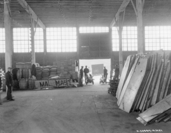 Man are standing in a factory building or warehouse, stacked with barrels, etc.
