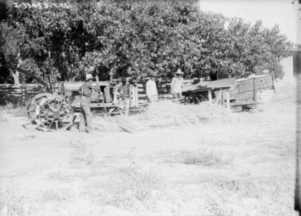 A group of men are working in a field with machinery being belt-driven with a Farmall tractor.