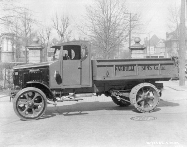 View across street towards a truck parked along the opposite curb. The sign on the side of the truck bed reads: "Nardulli & Sons Co. Inc." A park and houses are in the background.