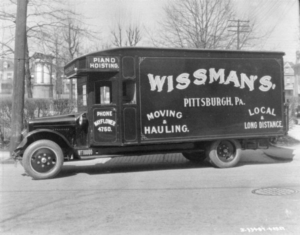 View across street towards a moving van parked along a curb. In the background is a park and houses. The sign painted on the side of the van reads: "Wissman's Moving & Hauling, Local & Long Distance," and "Piano Hoisting."