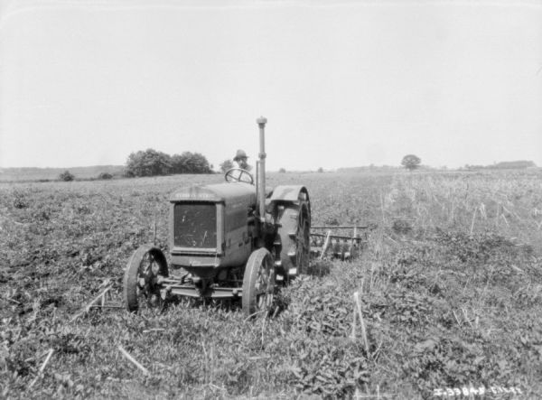 Three-quarter view from front left of a man driving a McCormick-Deering 15-30 tractor to pull a disk harrow in a field.