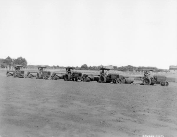 View across racetrack towards five men driving five industrial tractors pulling rollers to smooth out the track.